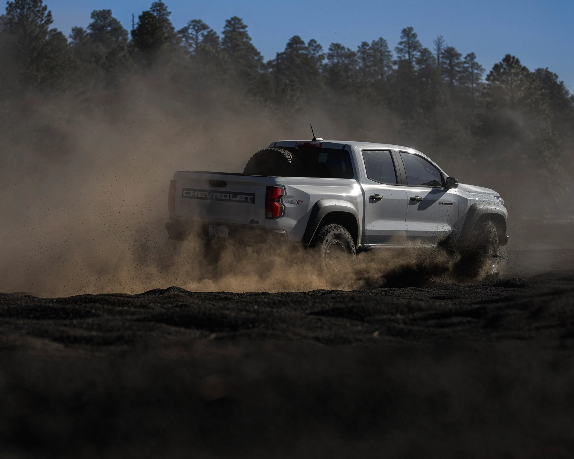 White Chevrolet pickup truck kicking up dust on a dirt road with trees in the background.