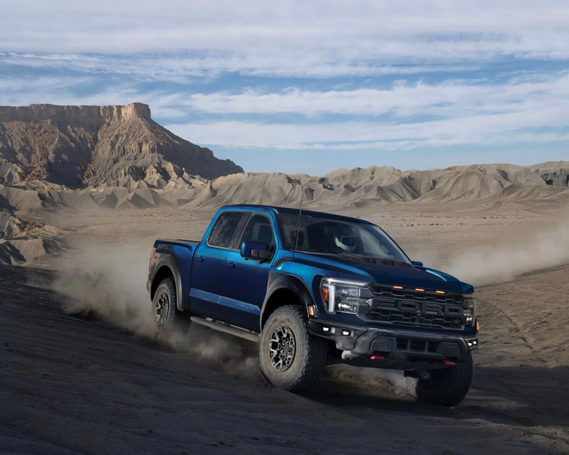 Blue Ford Raptor pickup truck driving on dusty desert terrain with rocky hills in the background.