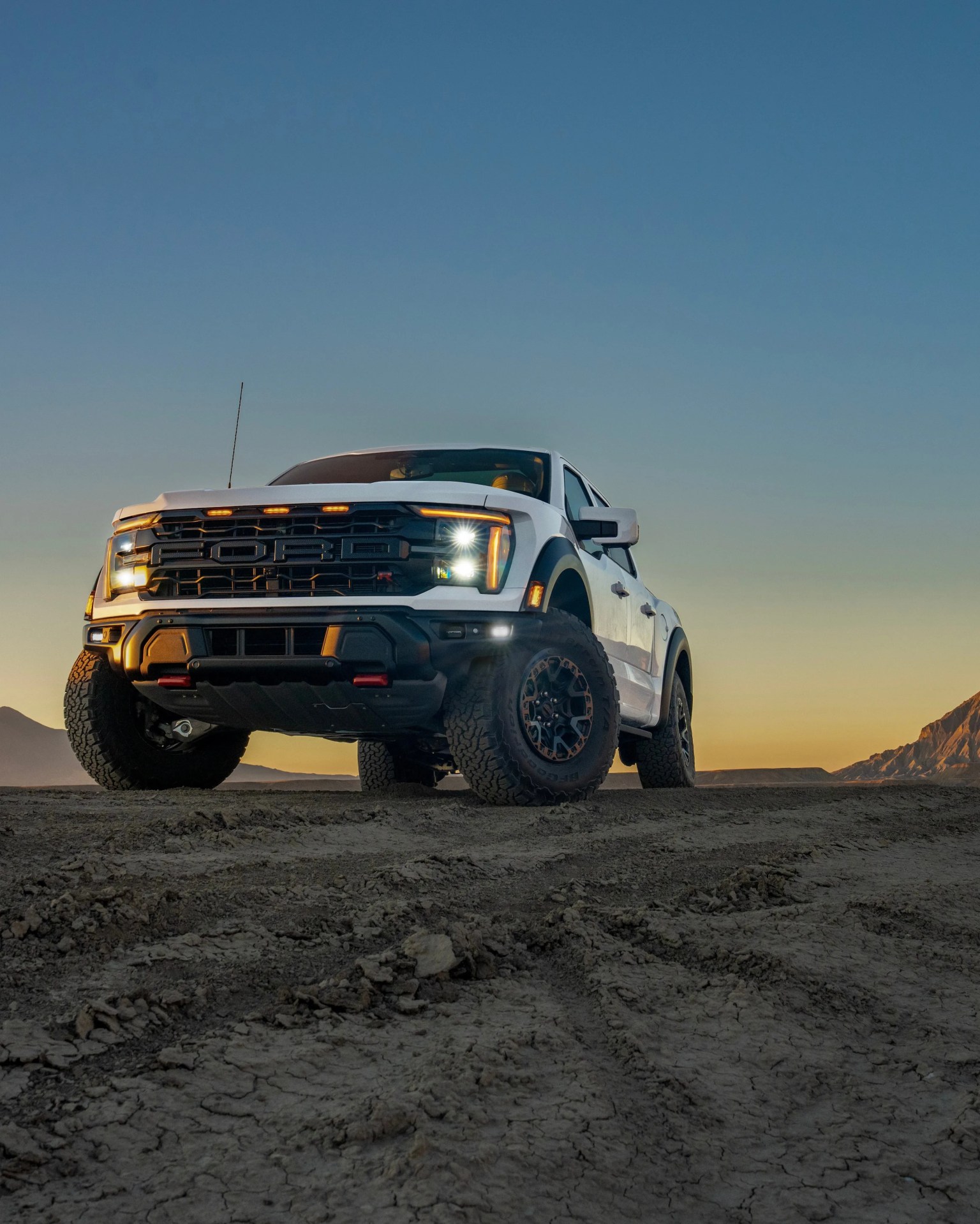 White Ford Raptor truck with headlights on parked on rough terrain at sunset.