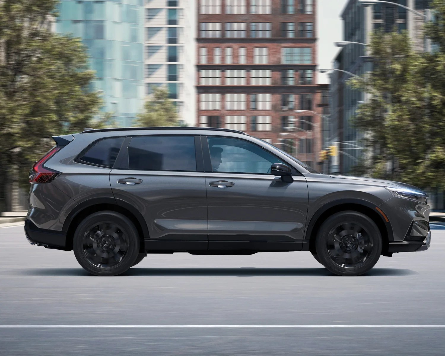 Gray mid-size SUV with black wheels driving on a city street with buildings and trees in the background.
