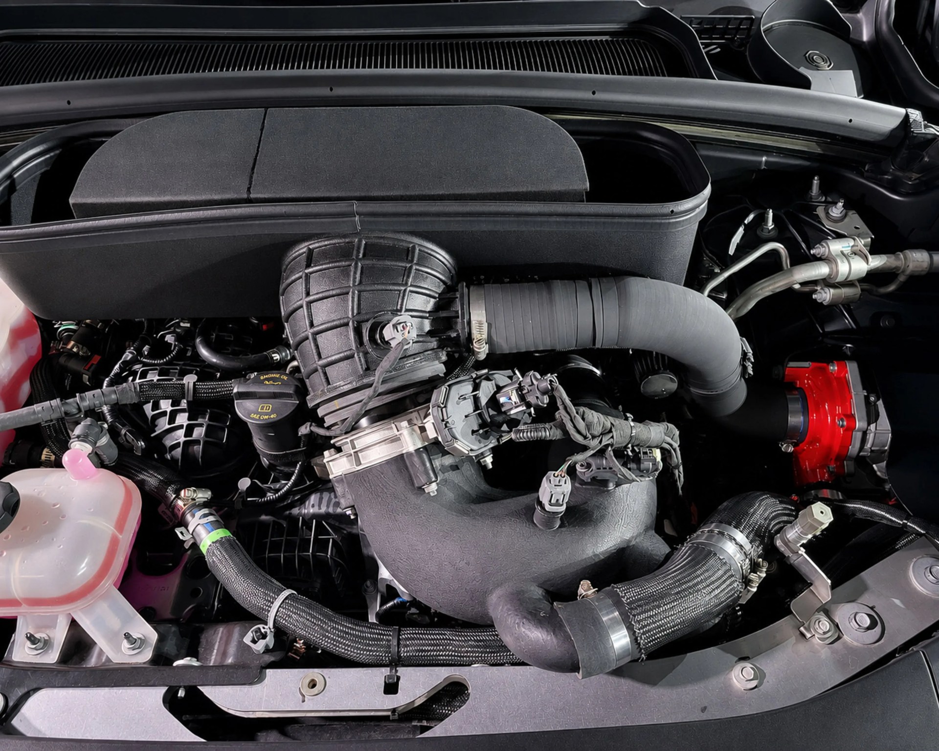 Close-up of a car engine compartment showing black air intake tubing, wiring, and a translucent coolant reservoir with pink fluid.