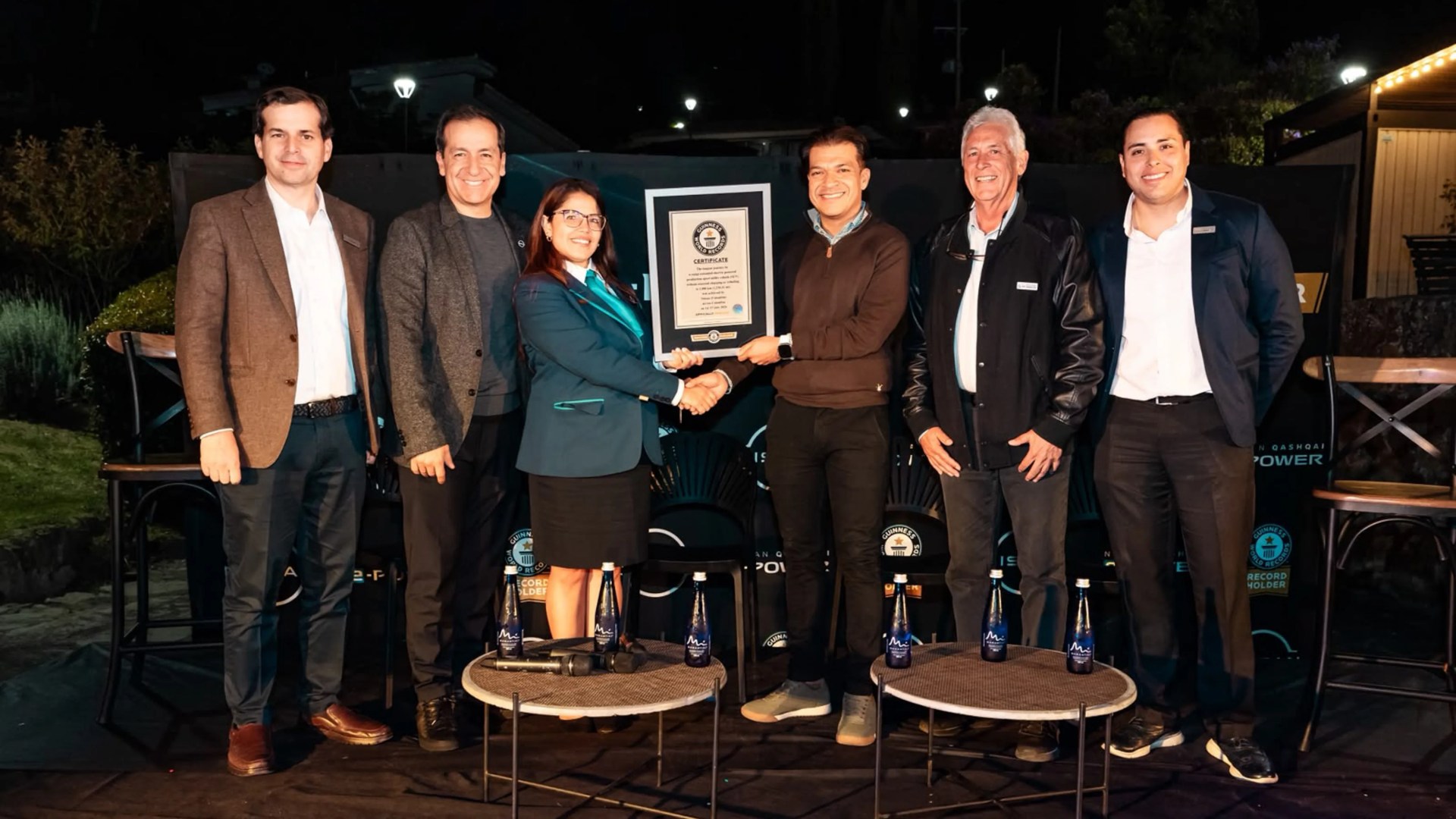 Six people standing in a row at night, two in the center shaking hands and holding a framed certificate, with small round tables and water bottles in front.