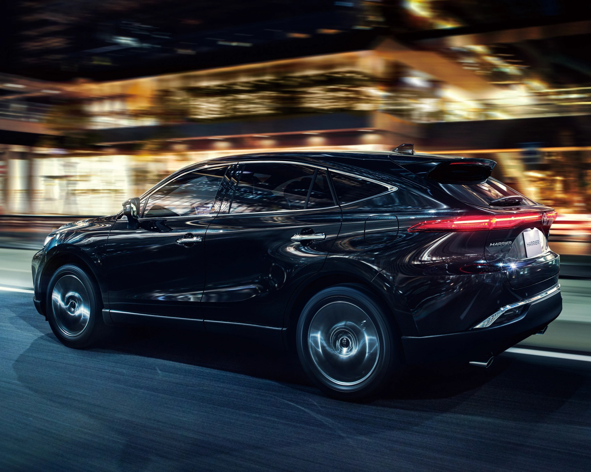 Black Toyota Harrier SUV driving on a city street at night with blurred lights in the background.
