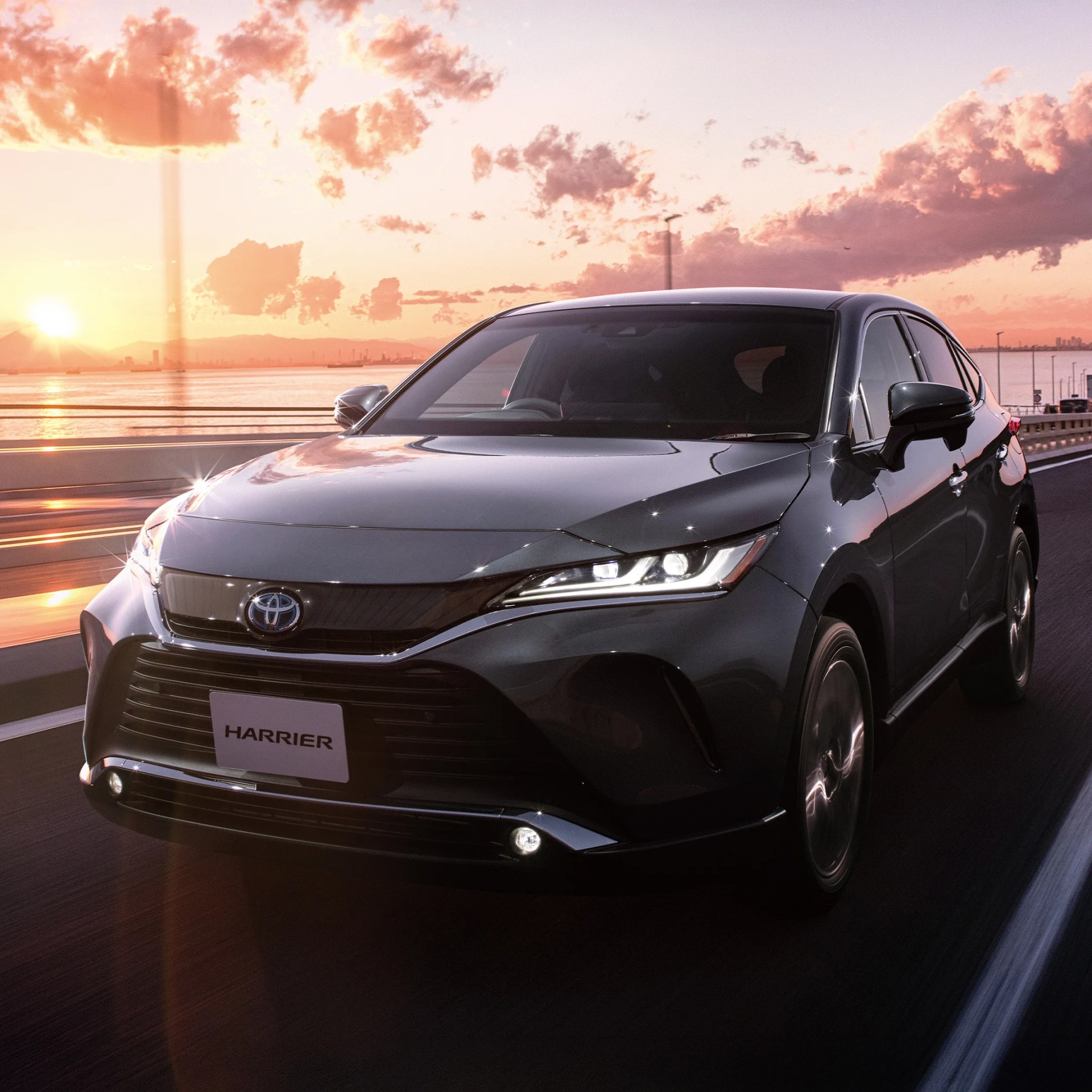 Black Toyota Harrier SUV driving on a road at sunset with a scenic sky and water in the background.