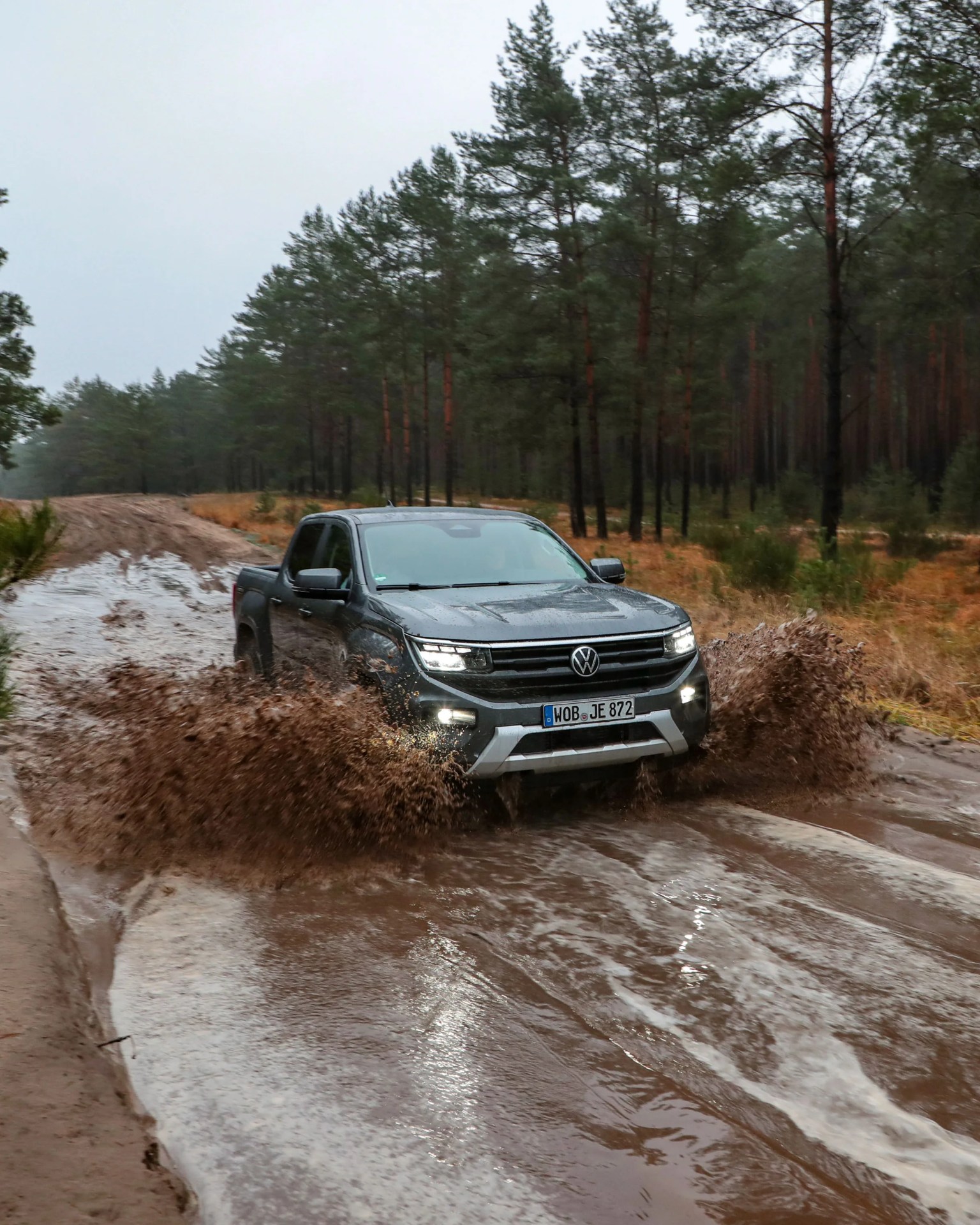 Gray Volkswagen pickup truck splashing through a muddy water puddle on a forest trail.