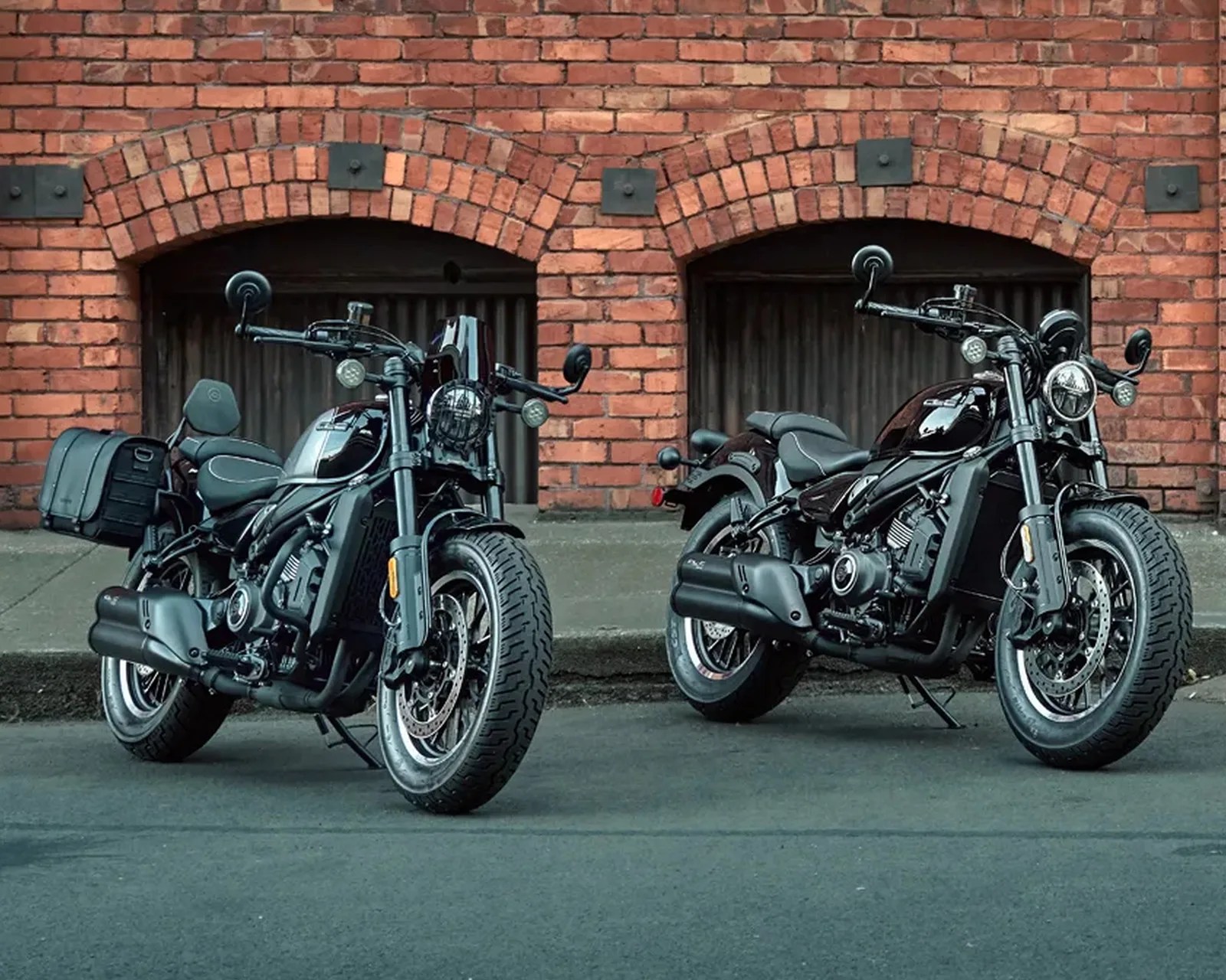 Two black cruiser motorcycles parked on a street in front of a red brick wall with arched doorways.