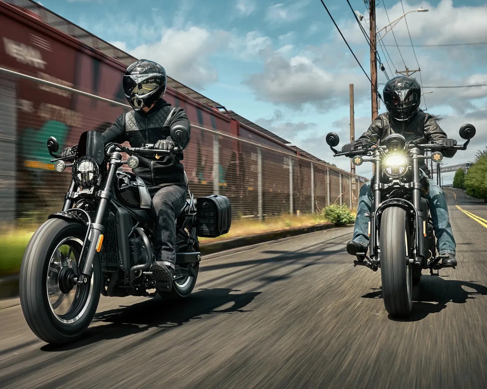 Two riders wearing black helmets and jackets on black motorcycles riding on a road beside a moving freight train.