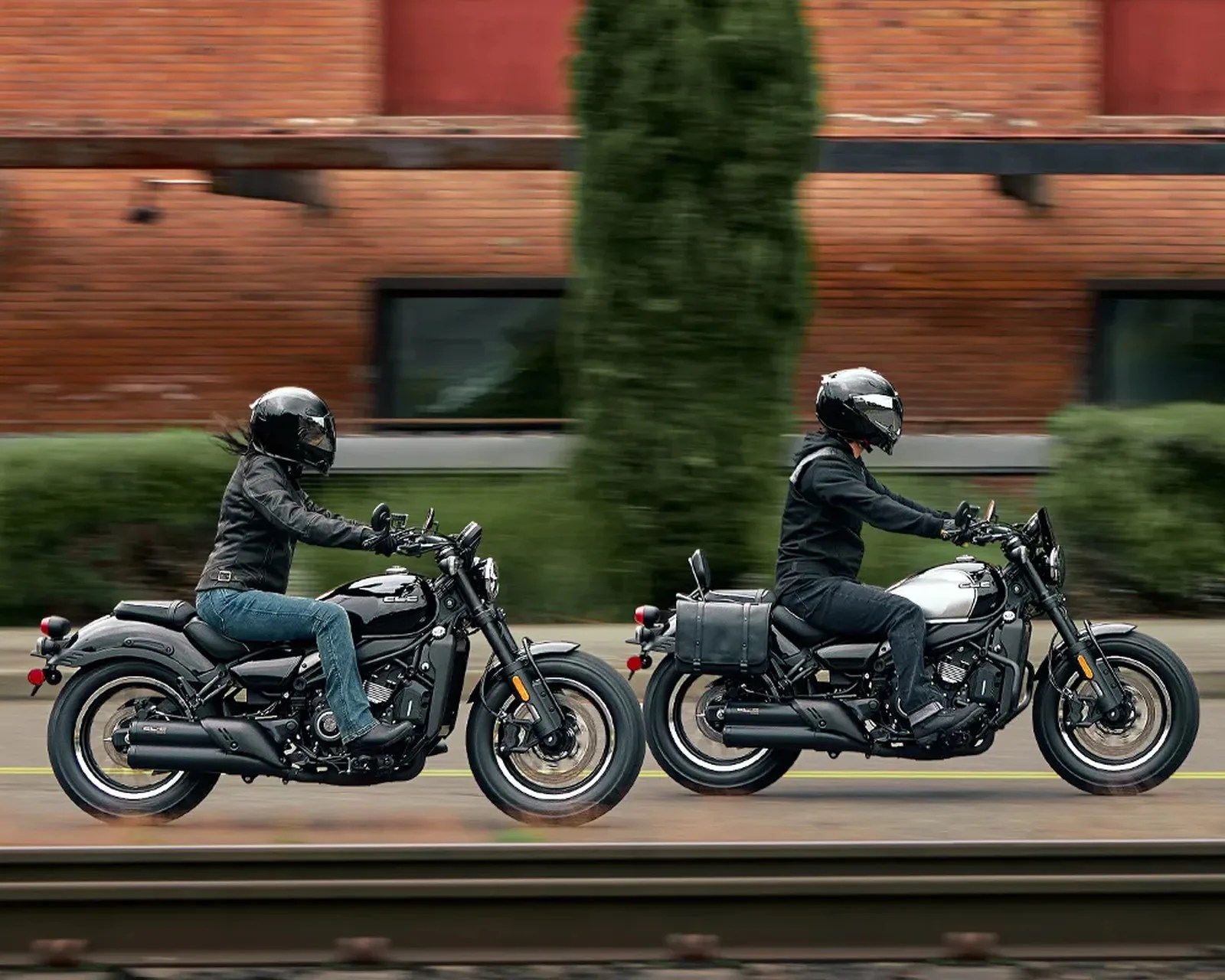 Two riders wearing black helmets and jackets riding black and silver motorcycles on a street with a brick building and greenery in the background.