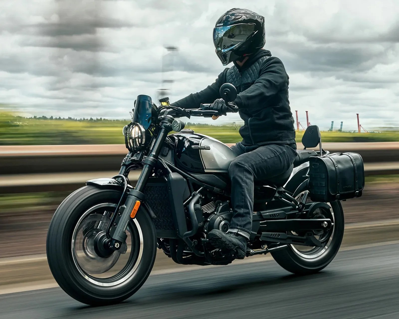 Person wearing black helmet and jacket riding a black and silver motorcycle with a side bag on a road.