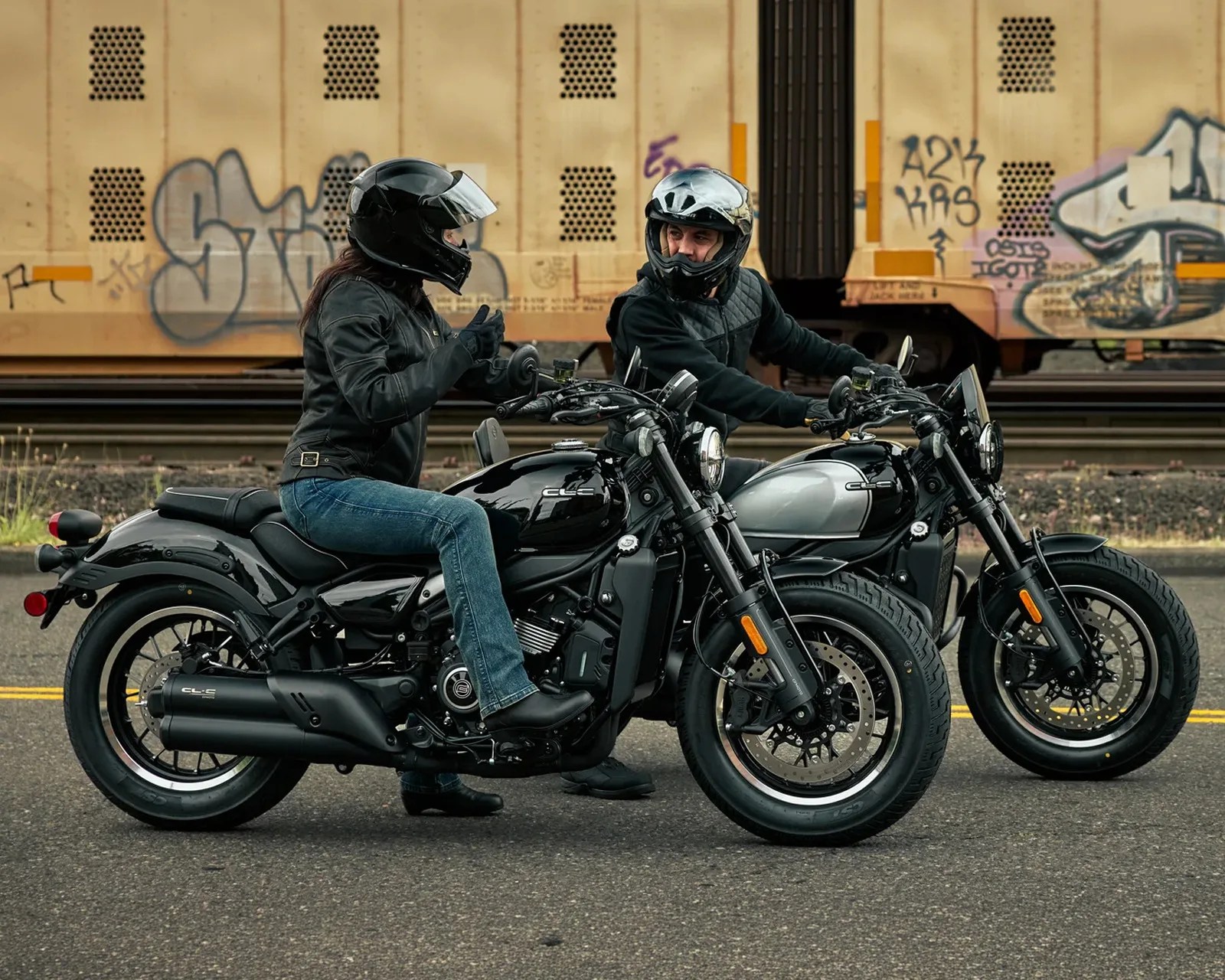 Two people wearing helmets and black jackets sitting on black and silver motorcycles on a street with a graffiti-covered train in the background.