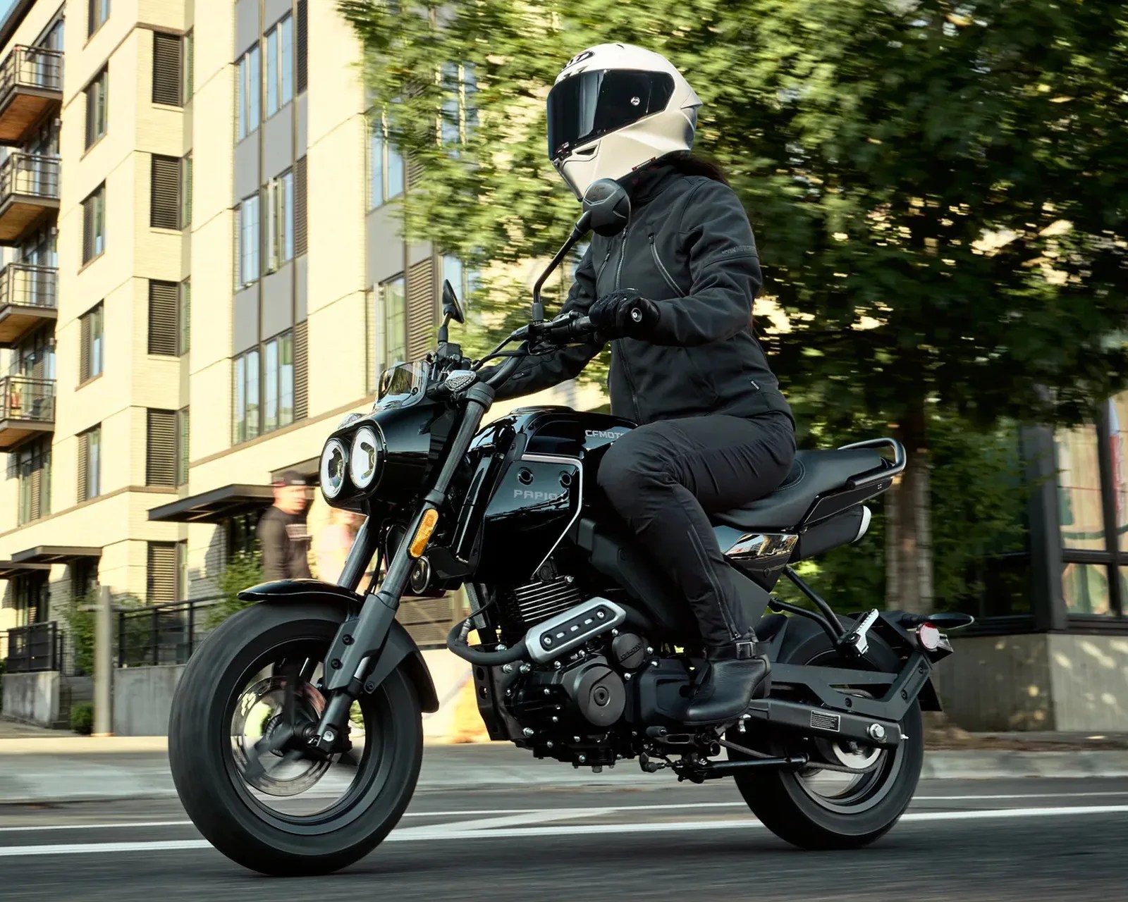 Person wearing a white helmet and black jacket riding a black CFMOTO motorcycle on a city street.