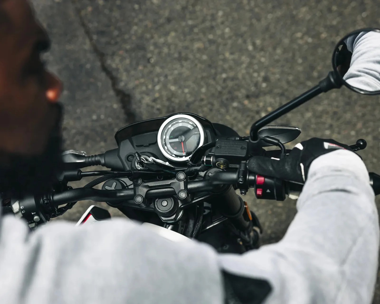Motorcycle dashboard with speedometer and rearview mirror, with rider wearing gloves and gray jacket.