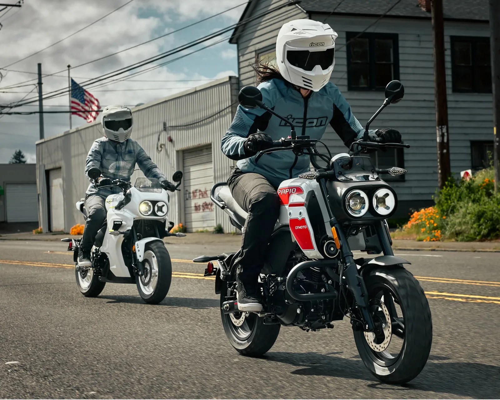 Two riders wearing white helmets and jackets riding white CFMOTO Papio motorcycles on a street with buildings and an American flag in the background.