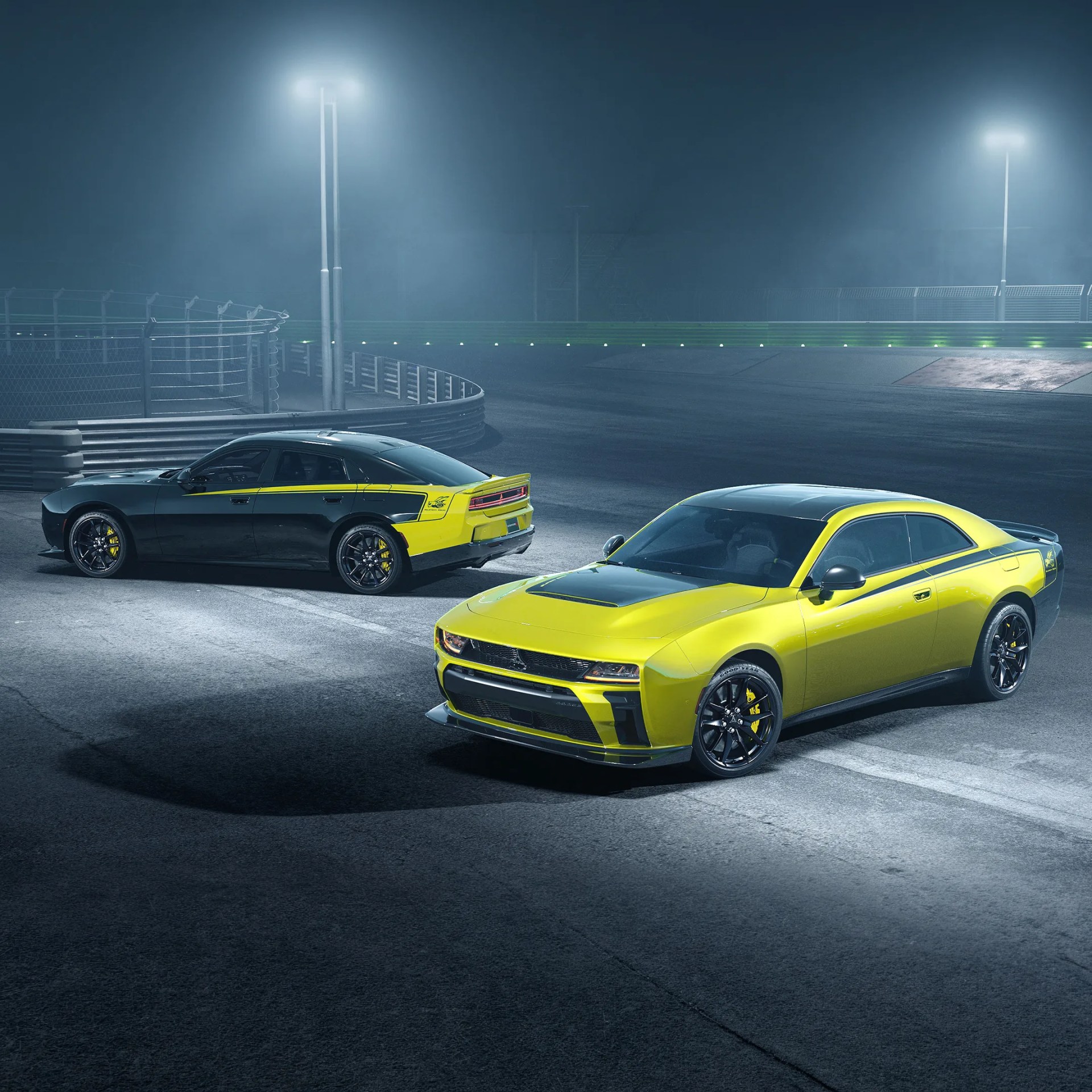 Two yellow and black Dodge muscle cars parked on a dimly lit racetrack at night.