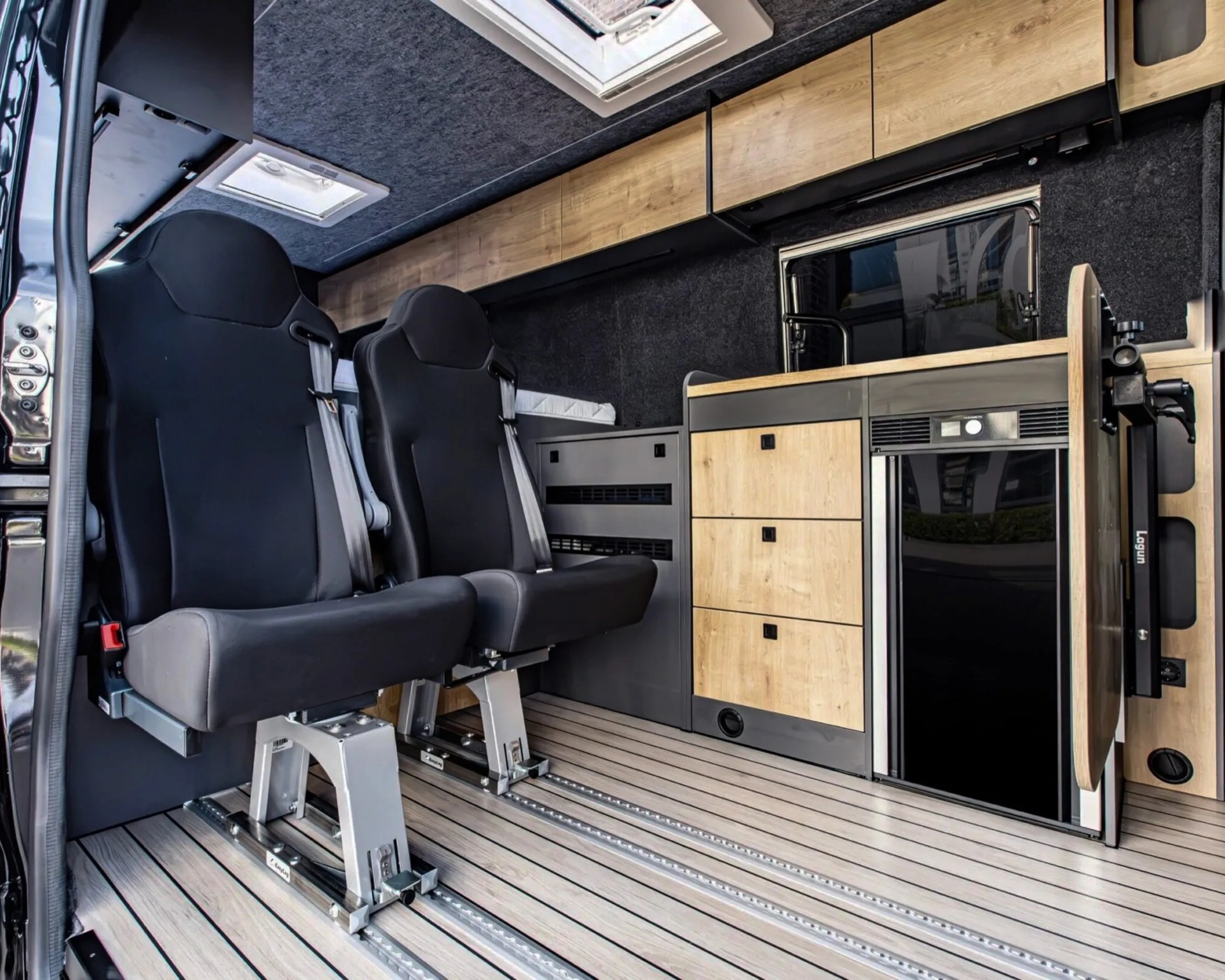 Two black swivel seats with seatbelts inside a camper van featuring wood-paneled cabinets and a small refrigerator.