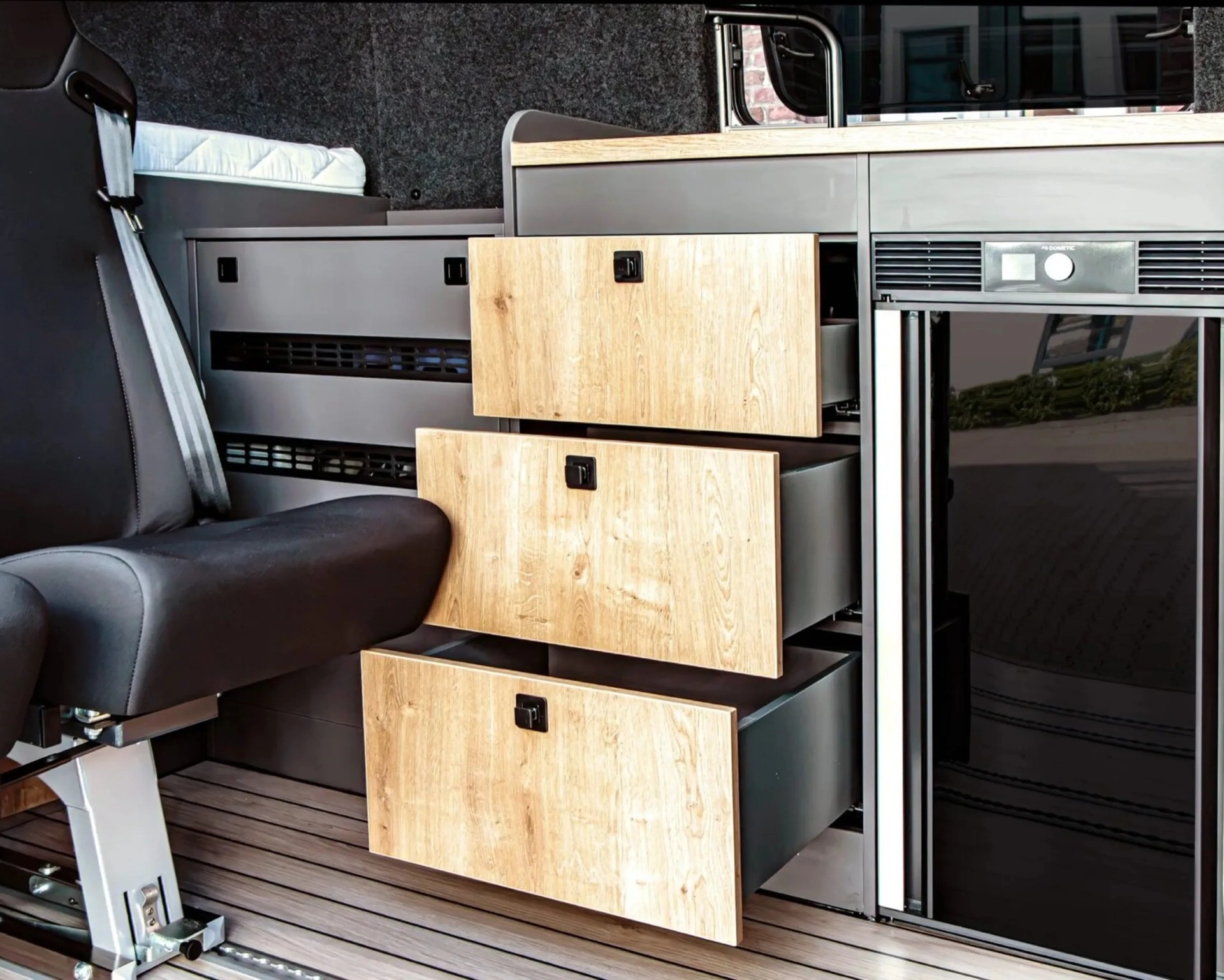 Black swivel seat with gray seatbelt next to open light wood drawers and a black mini fridge in a camper van interior.