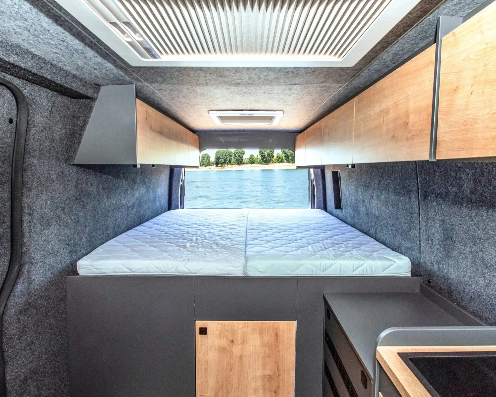 Interior view of a camper van with a white mattress, wood cabinets, gray walls, and a window showing water and trees outside.
