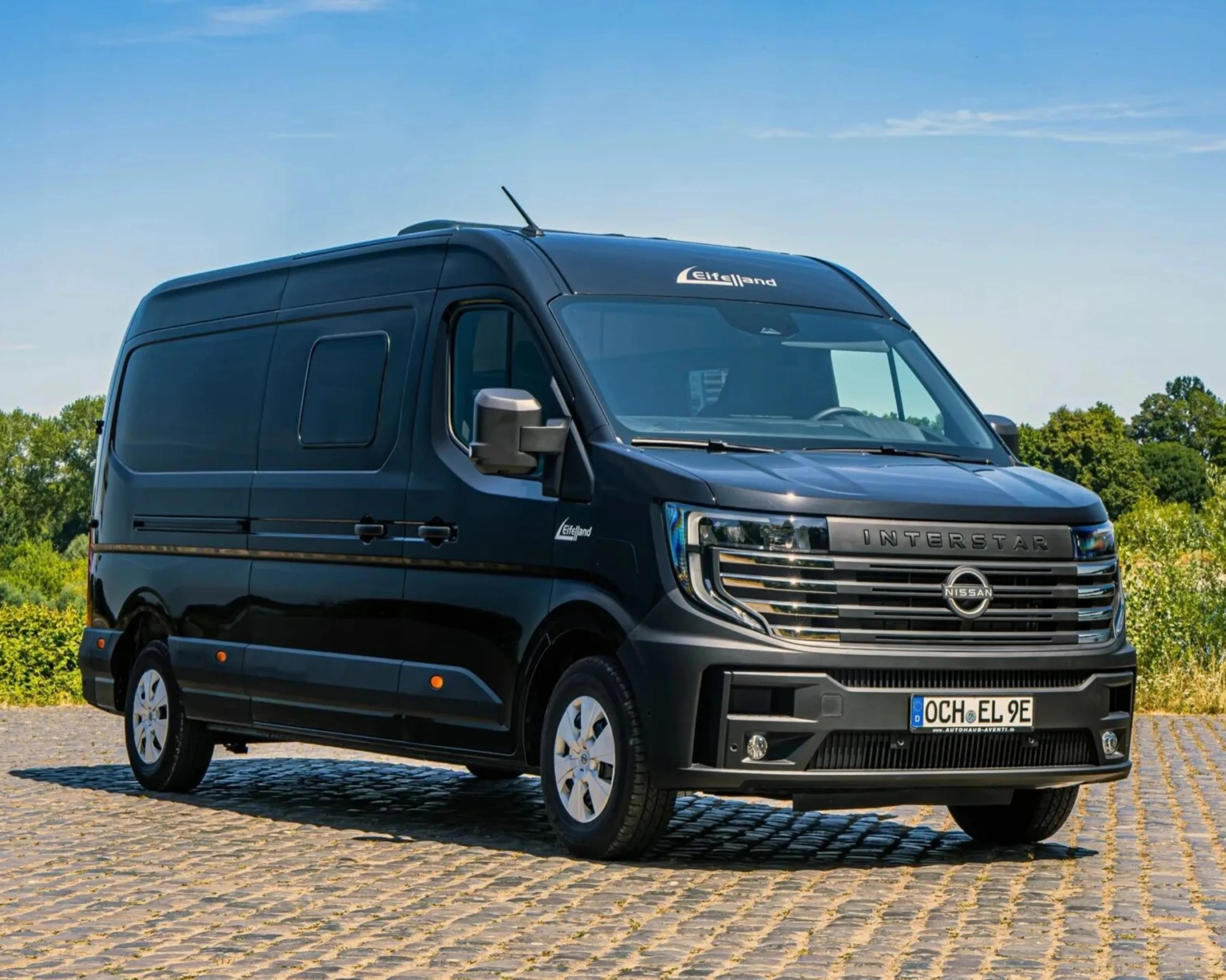 Black Nissan Interstar van with Eifelland branding parked on cobblestone pavement under a clear blue sky.