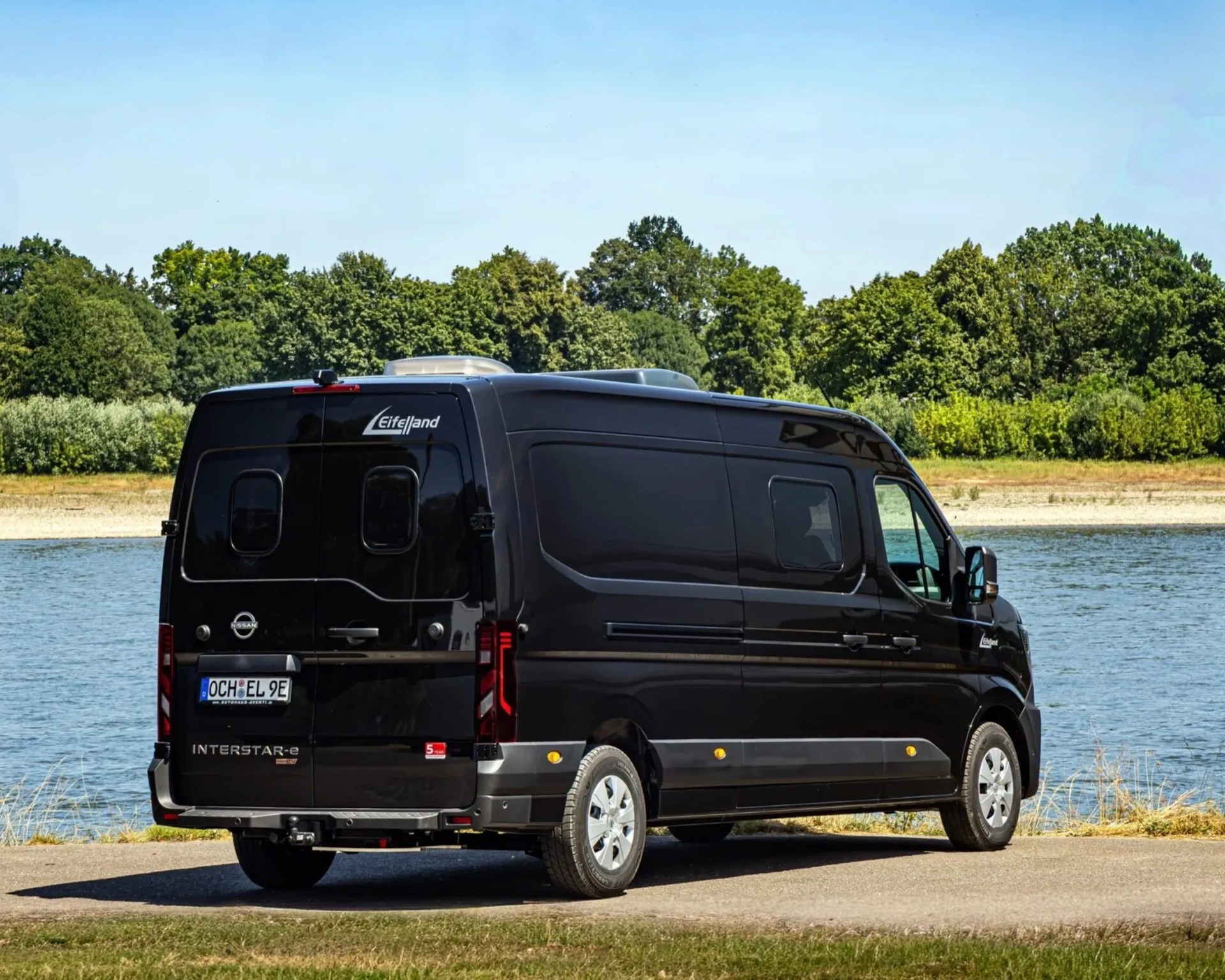 Black Nissan Interstar-e electric van parked near a river with green trees in the background.