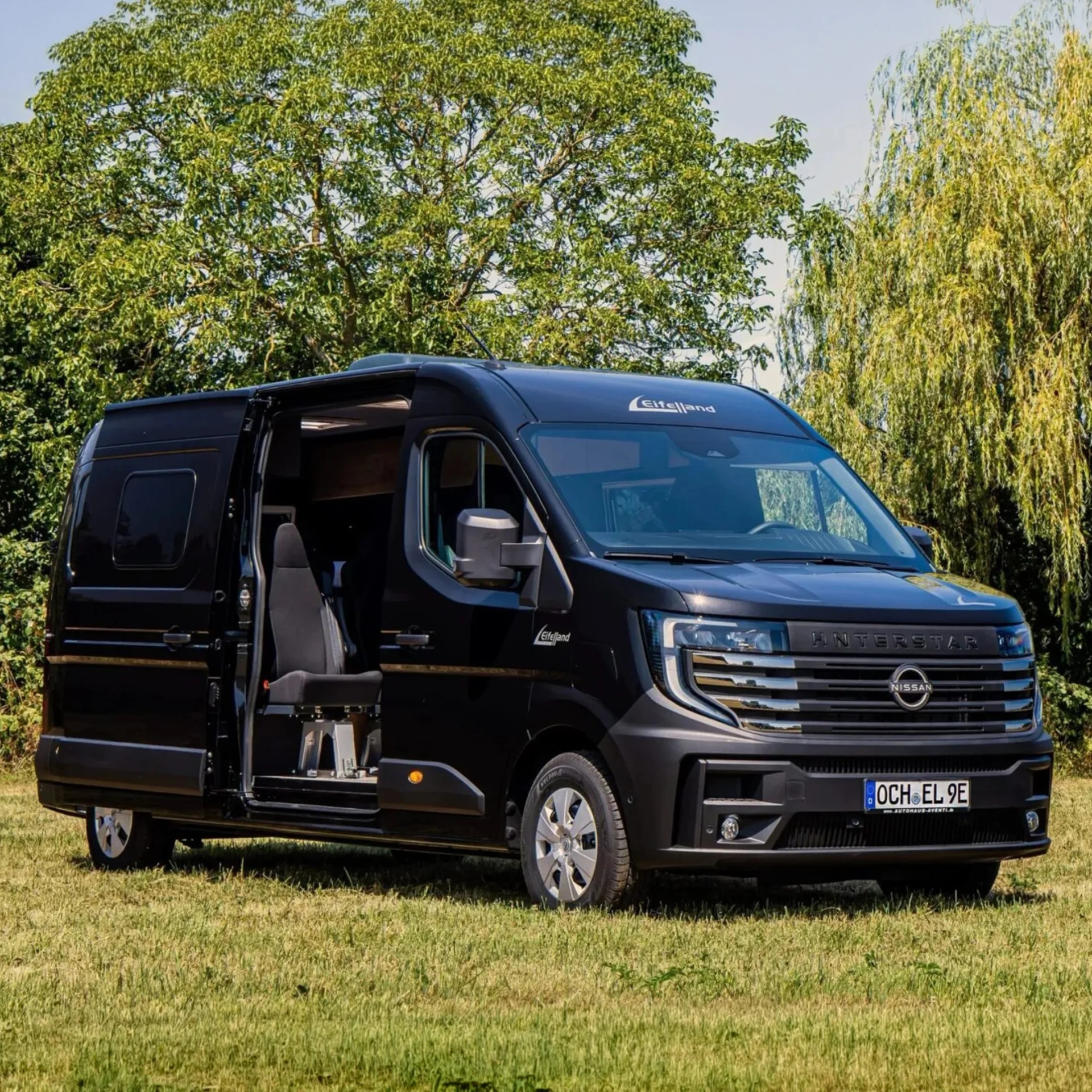 Black Nissan Interstar camper van with open sliding door and swivel seat on grass field with trees in background.