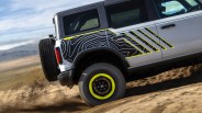 White off-road vehicle with black and yellow topographic pattern decal and neon yellow wheel rim driving on dirt.