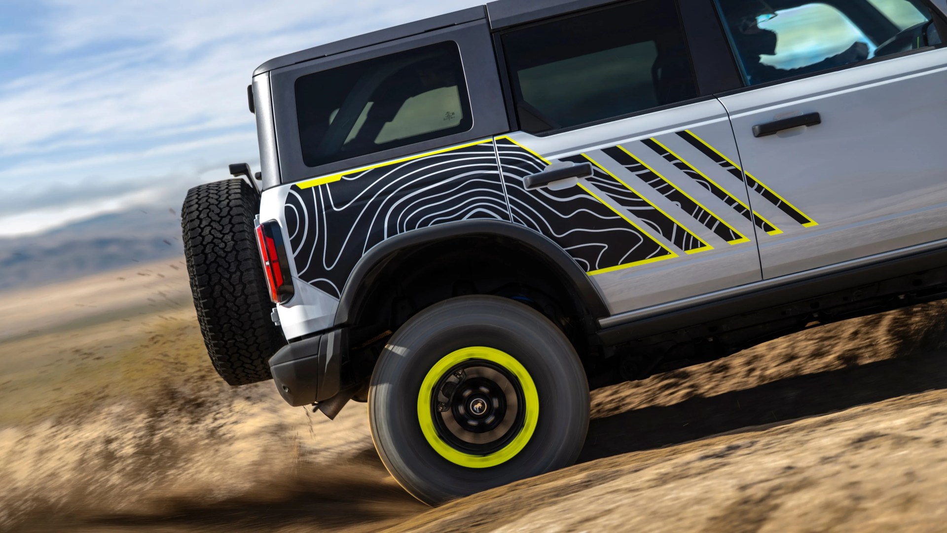 White off-road vehicle with black and yellow topographic pattern decal and neon yellow wheel rim driving on dirt.