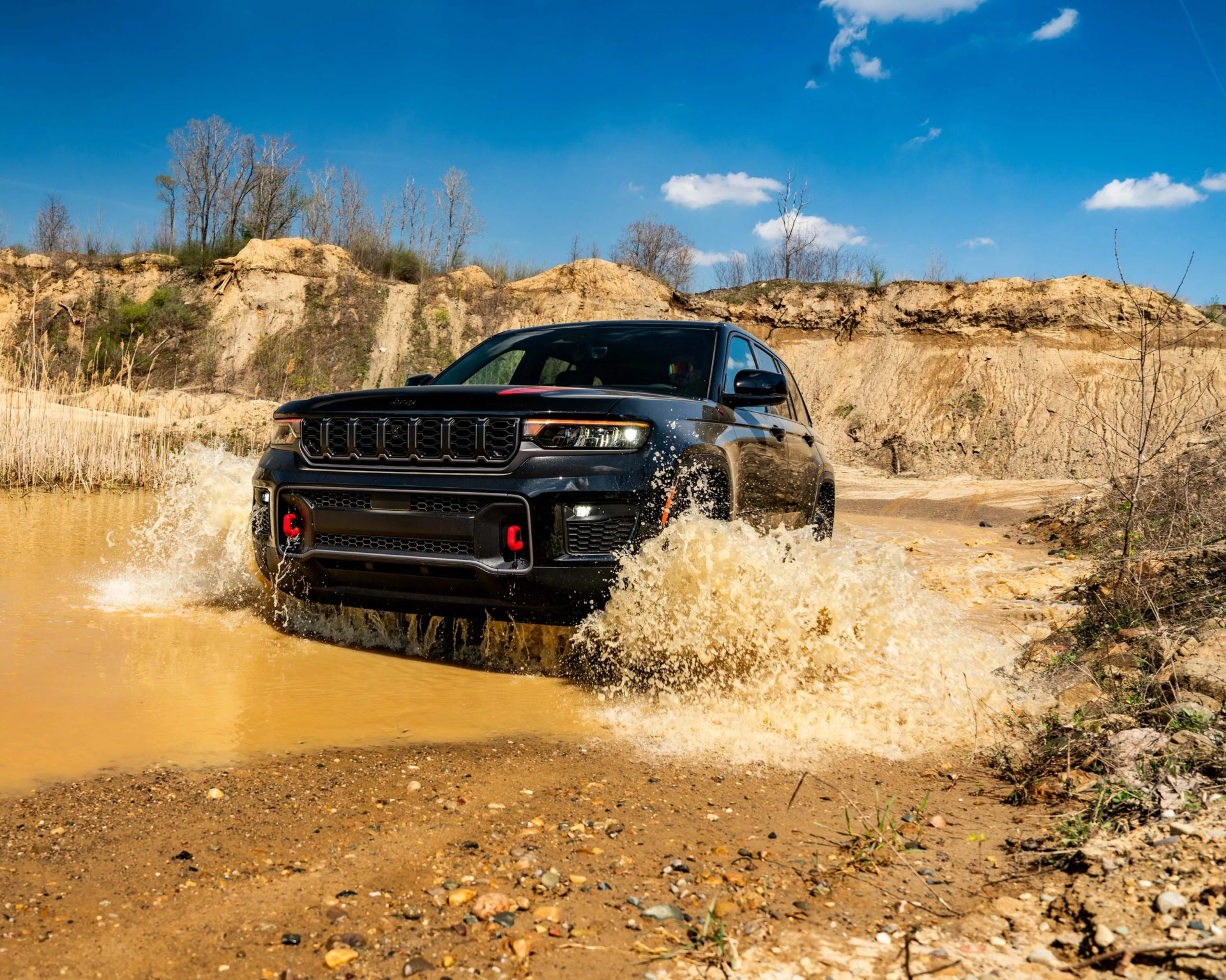 Black Jeep SUV splashing through muddy water in a rugged outdoor setting with dry trees and blue sky.
