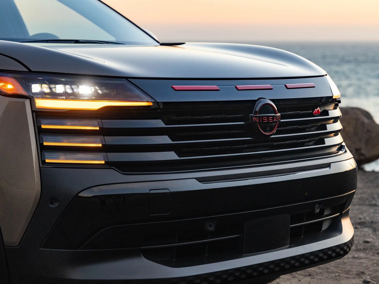 Front view of a black Nissan vehicle with illuminated headlights and amber turn signals near a body of water at sunset.