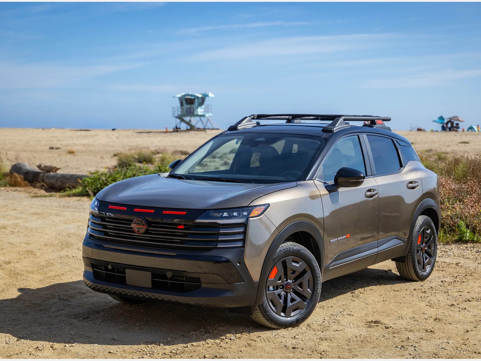 Metallic gray Nissan SUV with black roof and red accents parked on sandy terrain near a beach.