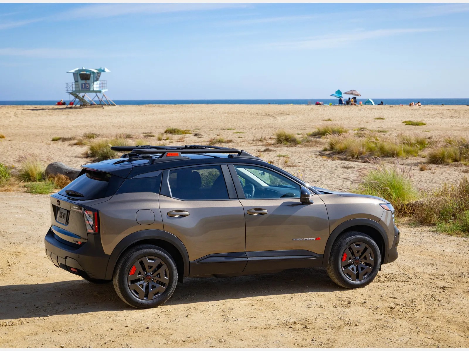 Gray and black compact SUV with roof rails parked on sandy beach near ocean and lifeguard tower.