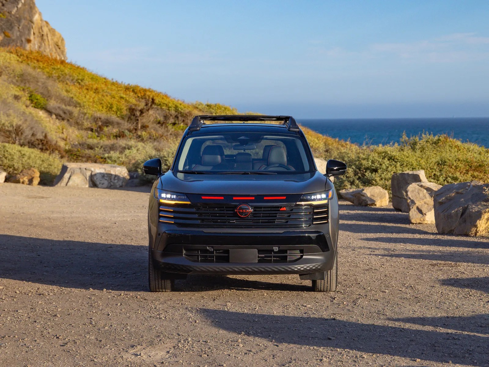Front view of a black Nissan SUV parked on a dirt road near rocky terrain and ocean.
