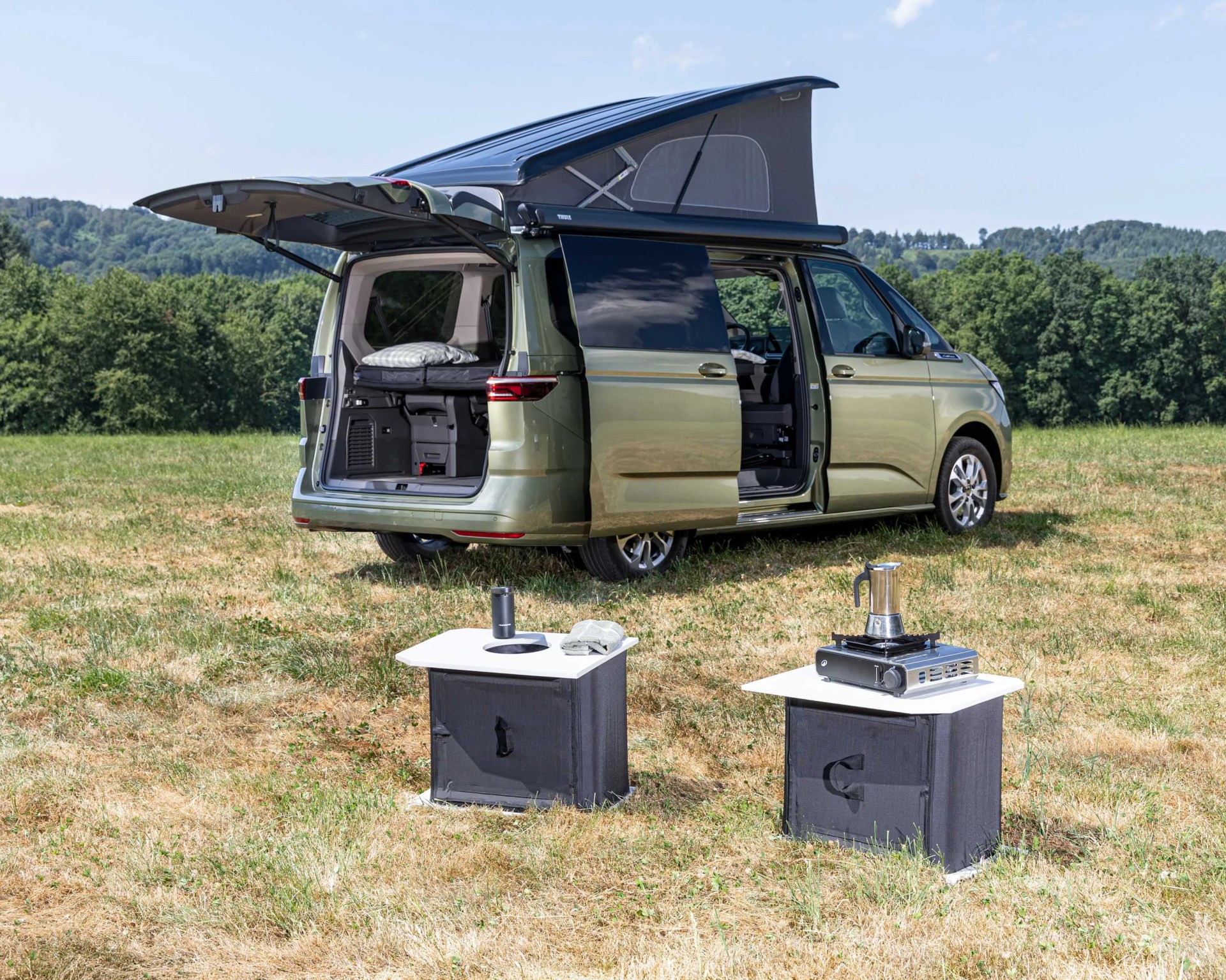 Green camper van with pop-up roof and open rear and side doors parked on grass with camping gear in front.