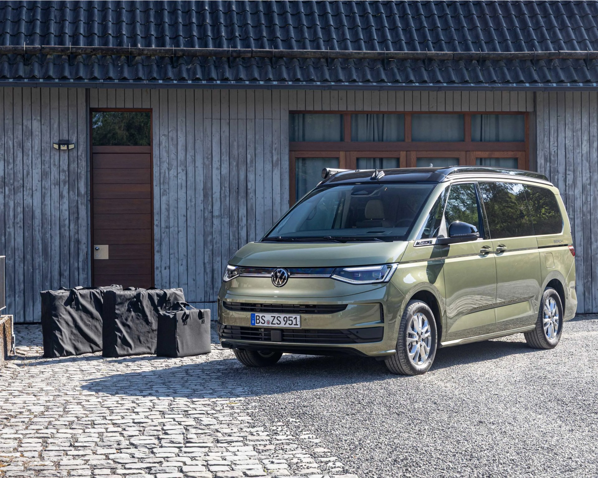 Green Volkswagen van parked on a cobblestone driveway in front of a wooden house with black roof tiles.