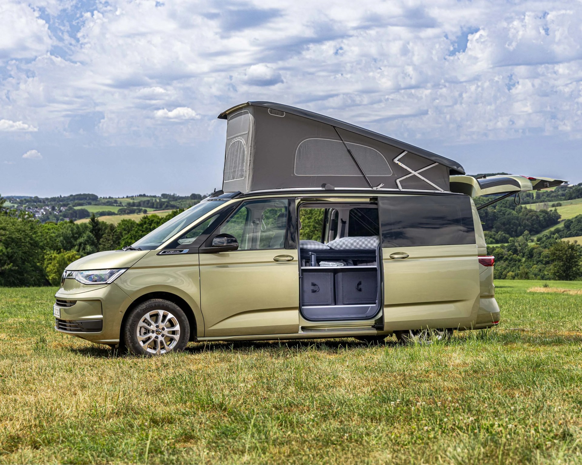 Light green camper van with pop-up roof and open sliding door parked on grassy field under cloudy sky.
