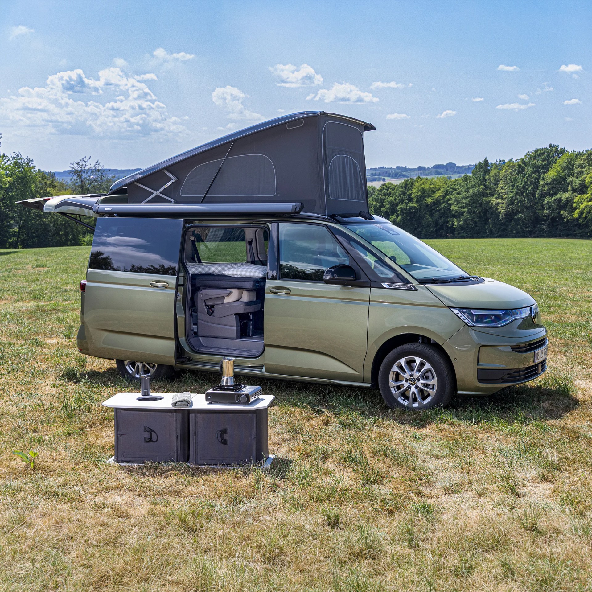 Green camper van with pop-up roof and open sliding door parked on grass with portable stove and table in front.