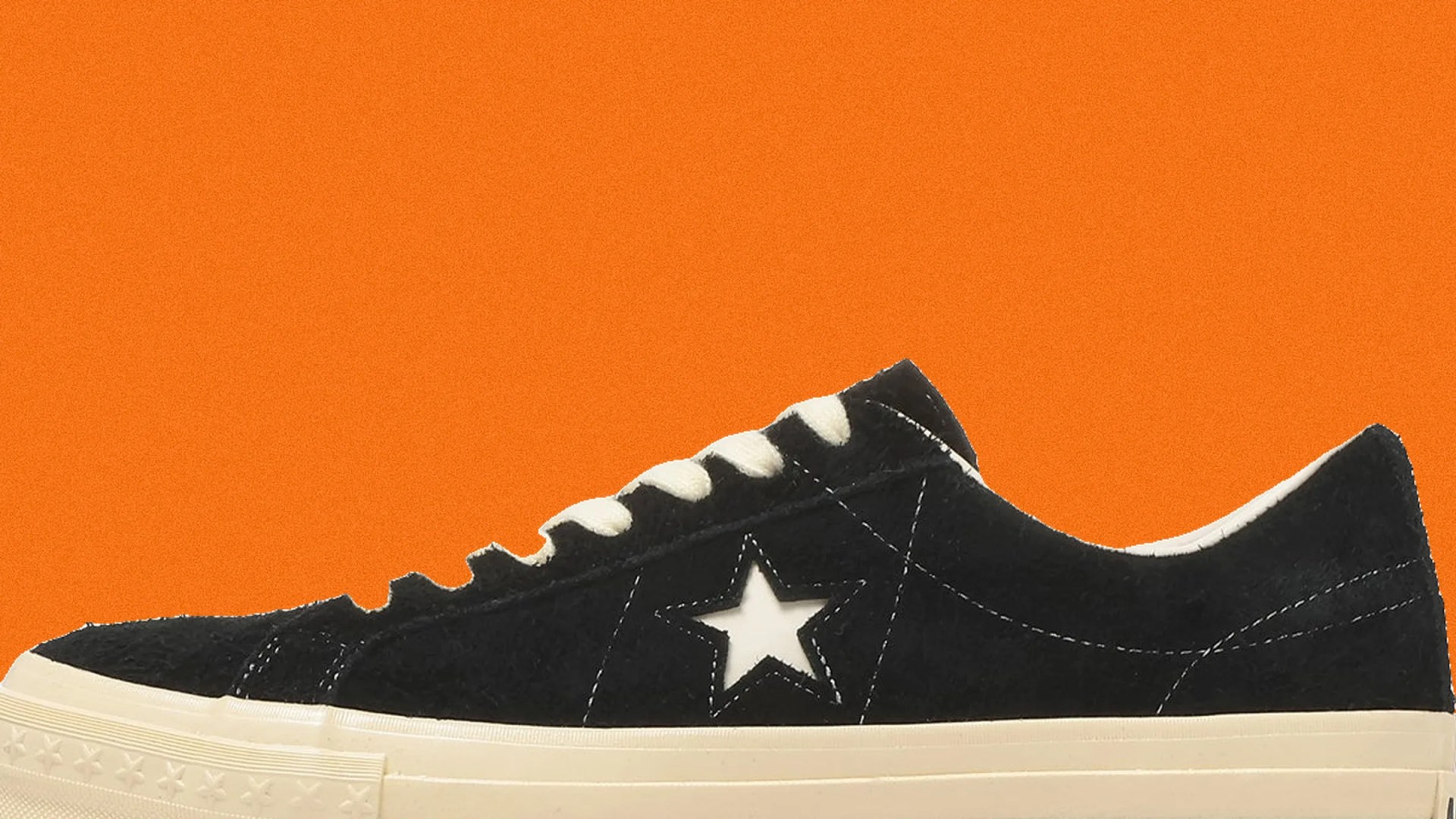 Black suede sneaker with white star logo and cream sole against an orange background.