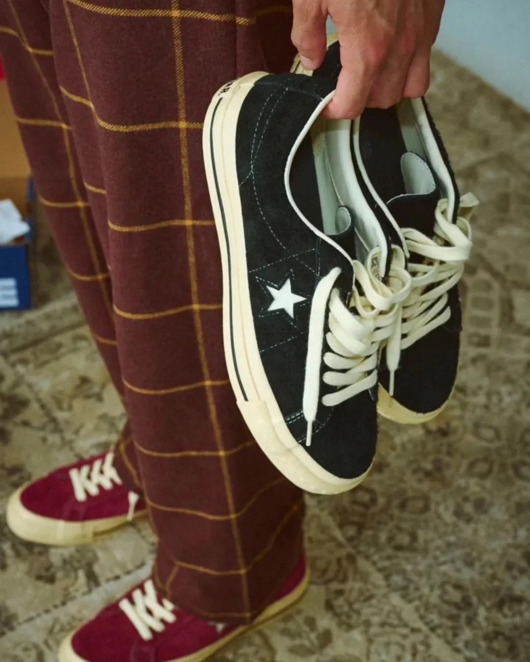 Person holding a pair of black Converse sneakers with white laces and star logo, wearing red Converse shoes and brown plaid pants.