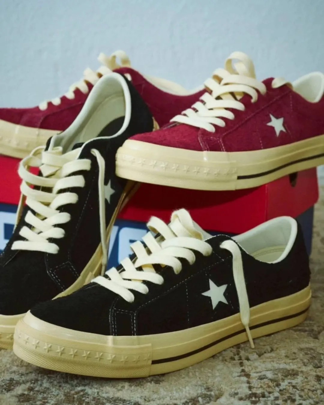 Two pairs of low-top Converse sneakers, one black and one burgundy, with cream laces and soles, displayed on a carpet.