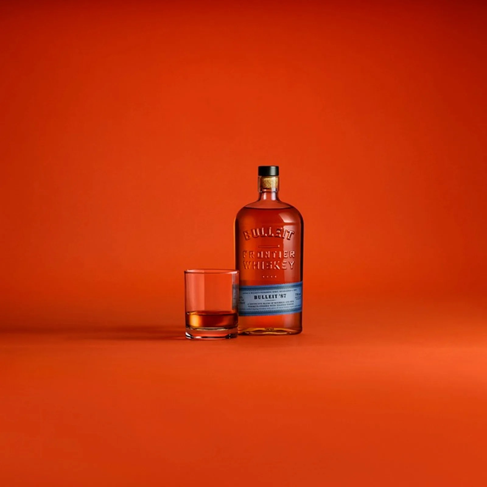 Bottle of Bulleit Frontier Whiskey with a filled glass on an orange background.