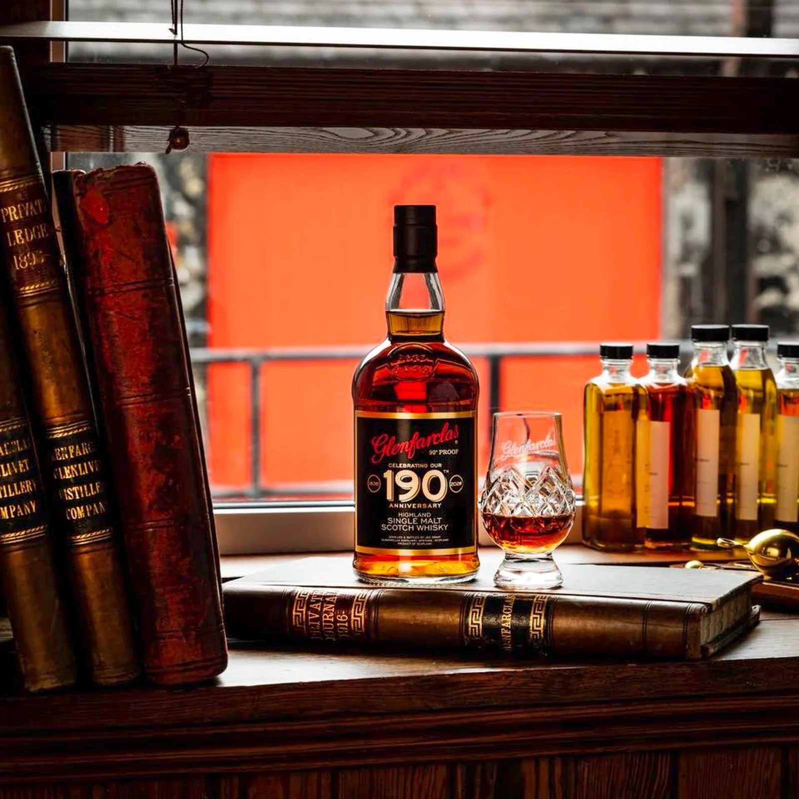 Bottle of Glenfarclas 190th Anniversary Highland Single Malt Scotch Whisky with a filled whisky glass on a windowsill surrounded by old books and small whisky bottles.