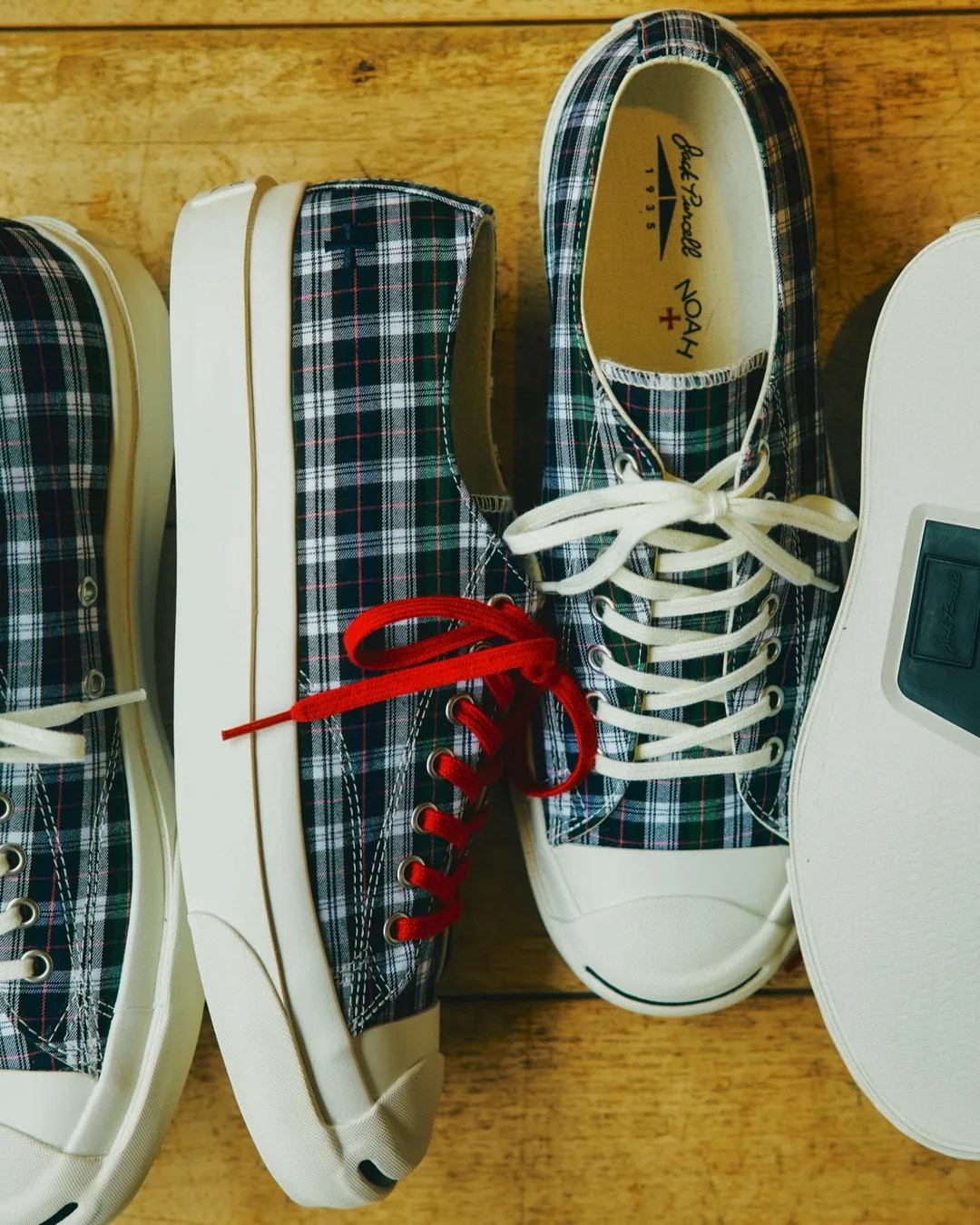 Pair of plaid sneakers with white rubber soles, one shoe with red laces and the other with white laces, on a wooden surface.