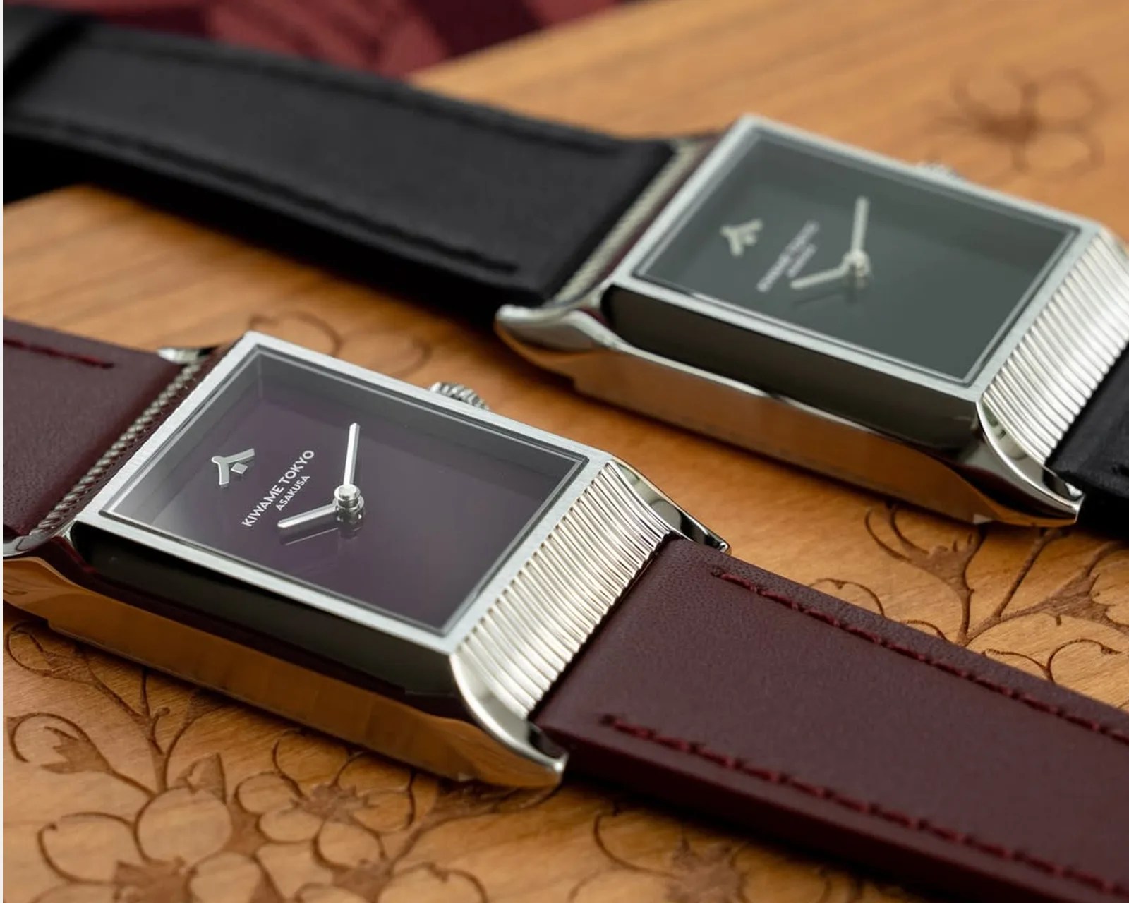 Two rectangular silver wristwatches with black and dark purple faces and leather straps on a wooden surface.