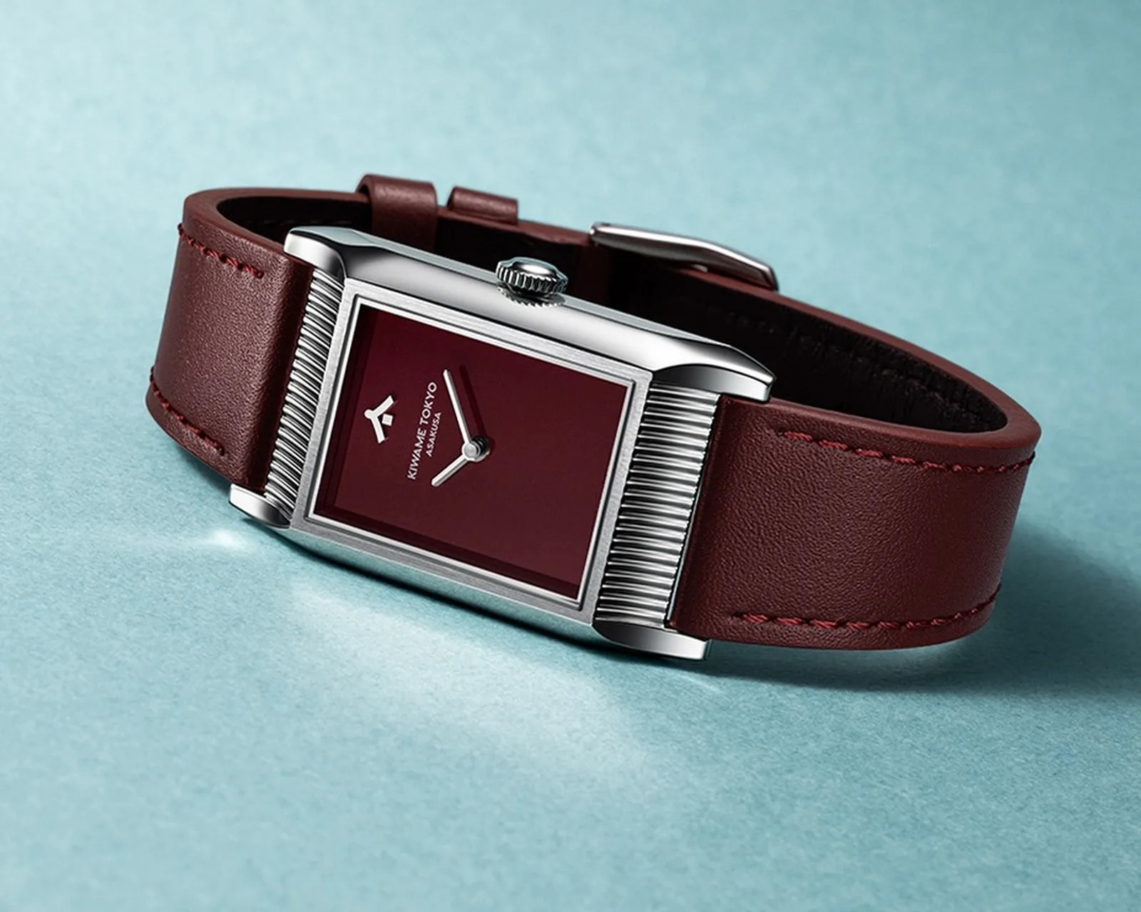 Rectangular silver watch with a burgundy dial and matching leather strap on a light blue surface.