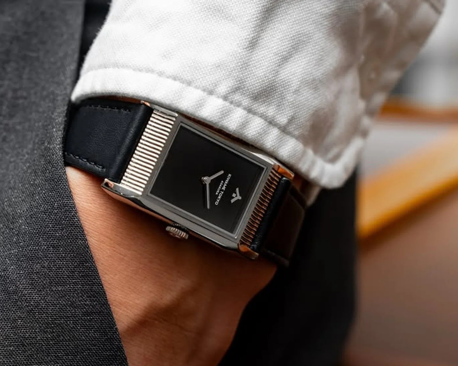 Rectangular wristwatch with black dial and silver ridged bezel on a black leather strap worn under a white cuff.