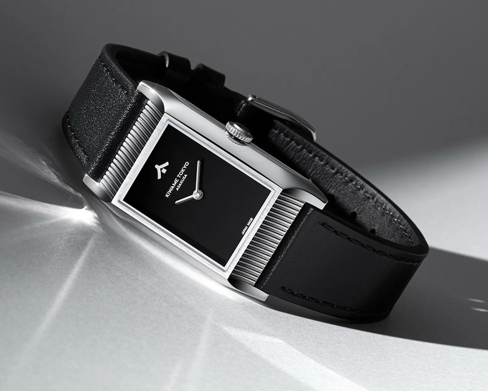 Rectangular silver watch with black dial and black leather strap on a gray surface.