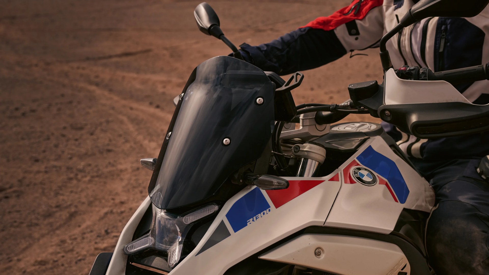 Close-up of a white BMW R1300 motorcycle with blue and red accents and a tinted windshield on a dirt surface.