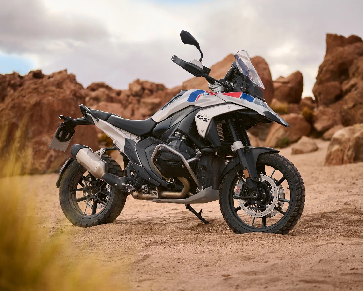 White and black BMW GS adventure motorcycle with knobby tires parked on sandy terrain with rocky background.