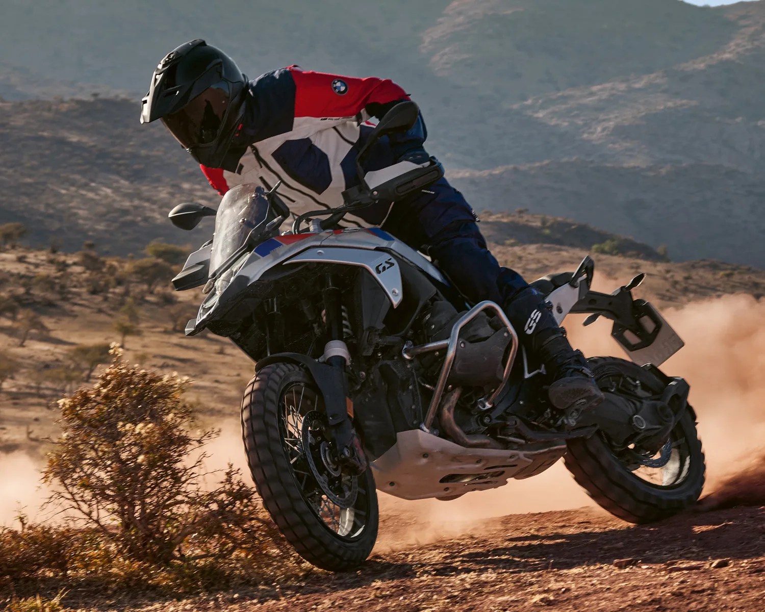 Motorcyclist in black helmet and red, white, and blue jacket riding a BMW GS adventure bike on a dusty trail.