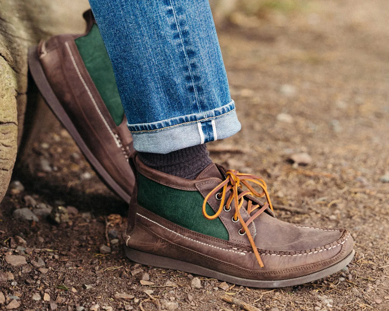 Brown and green leather moccasin-style shoes with orange laces worn with cuffed blue jeans on a dirt ground.