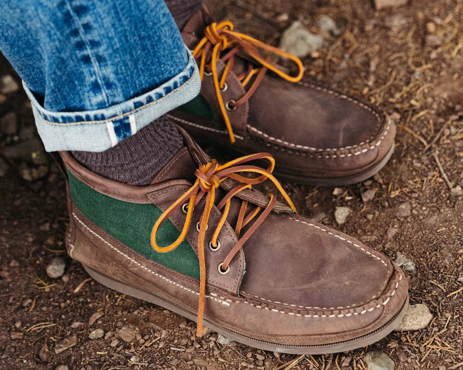 Brown and green leather moccasin boots with orange laces worn with cuffed blue jeans on dirt ground.